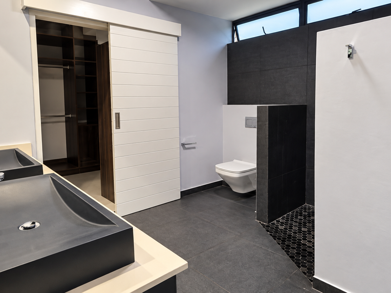 Modern bathroom featuring a double vanity with vessel sinks, wall-mounted toilet, dark floor tiling, and an integrated walk-in closet with sliding barn door access.