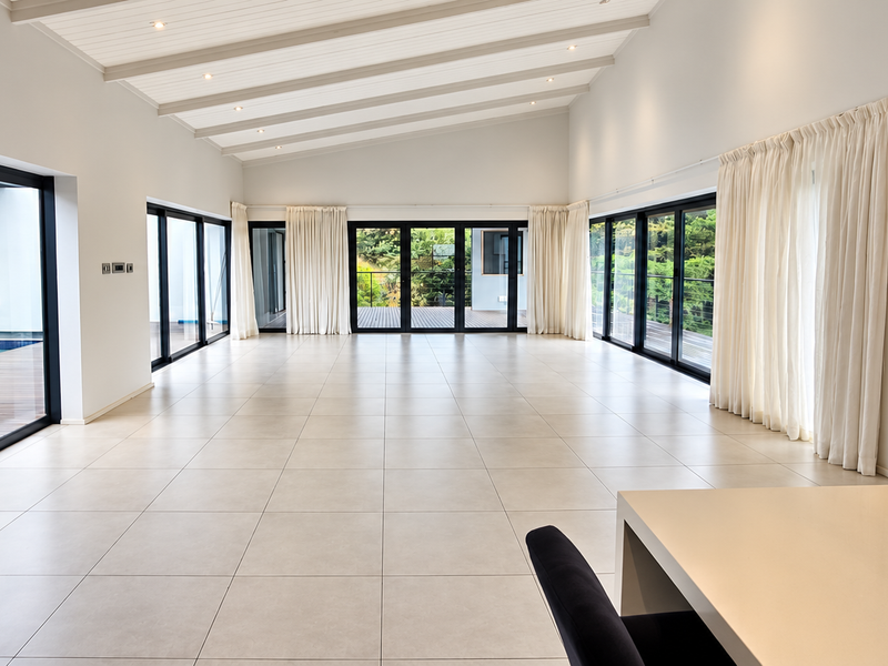 Spacious open-plan living area featuring large sliding glass doors, tiled flooring, and an exposed beam ceiling with recessed lighting, overlooking the pool area.