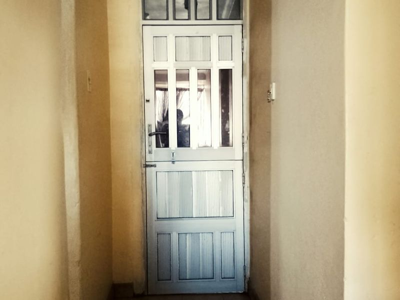 A narrow hallway featuring tiled flooring, a panelled door with a transom window, and coving, showing signs of wear on the wall surfaces.