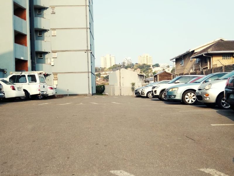An outdoor parking area situated at the rear of a multi-story apartment building with paved bays and perimeter fencing.