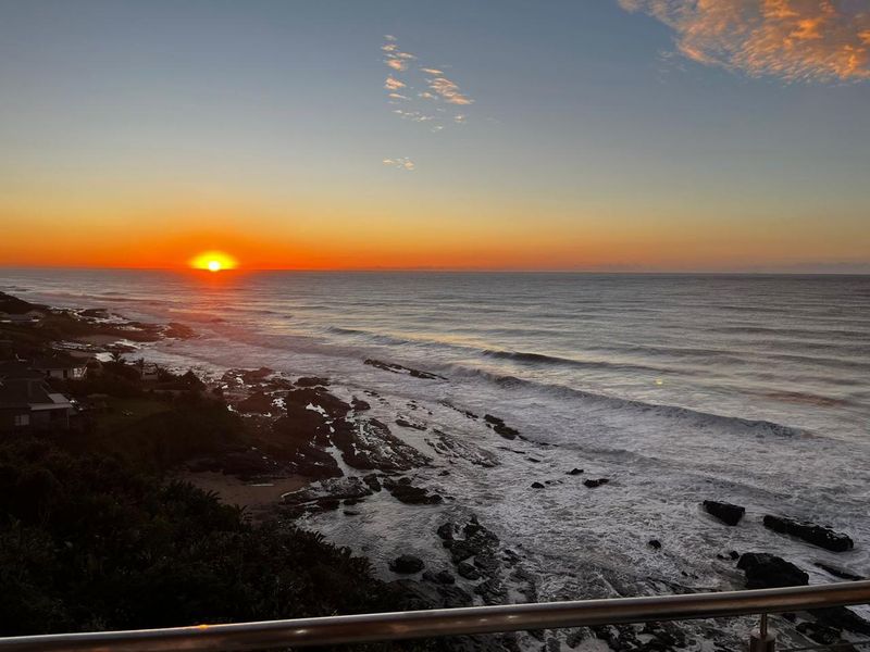 A stunning elevated ocean view featuring a sunset over the coastline, captured from a balcony with a modern metal railing.