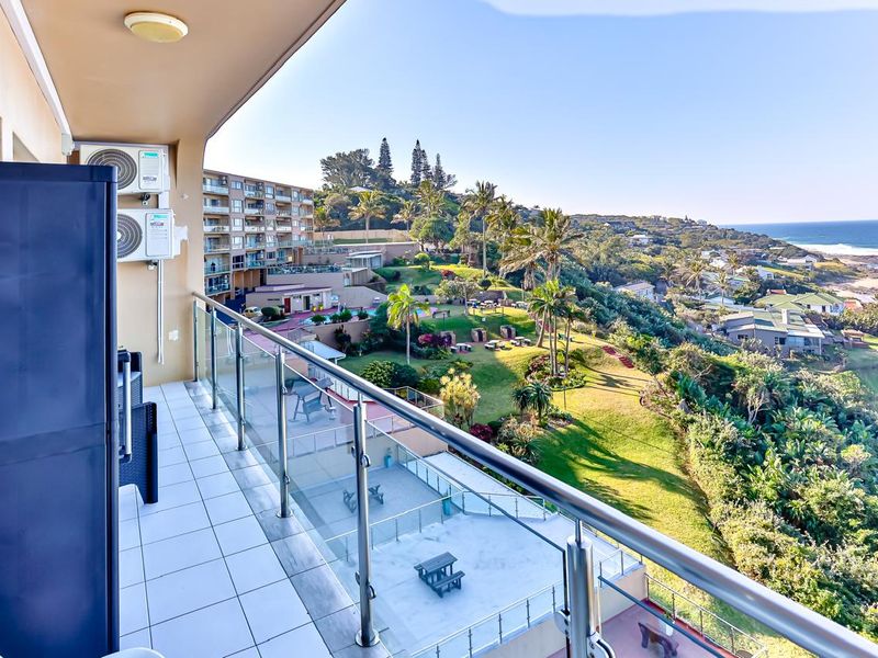 A spacious balcony featuring a modern glass balustrade, tiled flooring, and wall-mounted air conditioning units, offering expansive views of the coastal landscape and gardens.