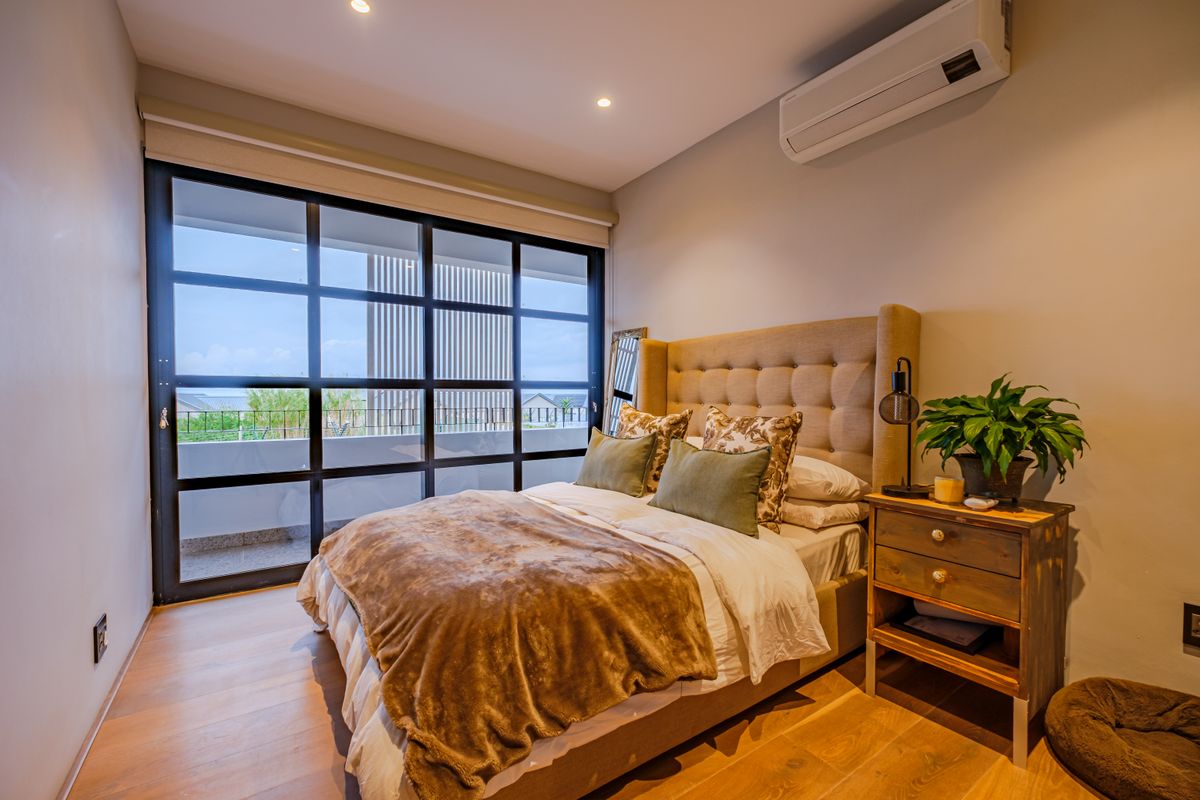 A well-maintained bedroom featuring timber flooring, recessed lighting, a wall-mounted air conditioner, and large glass sliding doors opening to a balcony.