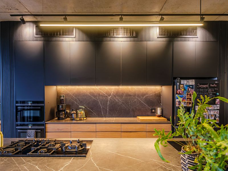 Modern kitchen featuring dark cabinetry, stone countertops, marble-look splashback, built-in appliances, and industrial-style concrete ceiling with integrated track lighting.
