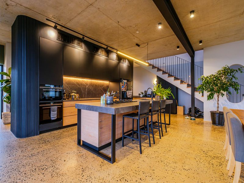 Modern open-plan kitchen featuring a large island, sleek cabinetry, exposed concrete ceilings, and polished concrete floors, seamlessly integrated with a staircase.