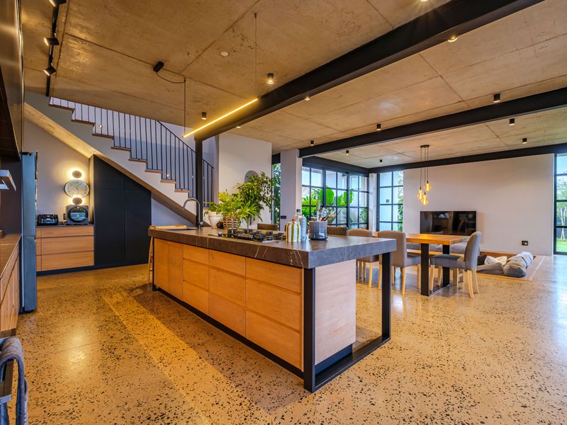 Modern open-plan living area featuring a sleek kitchen island, exposed concrete ceilings, steel structural elements, and polished concrete floors with seamless indoor-outdoor flow.