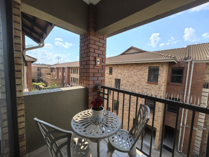 A covered balcony featuring brick pillars, metal balustrading with security spikes, and tiled flooring, overlooking the surrounding townhouse complex.