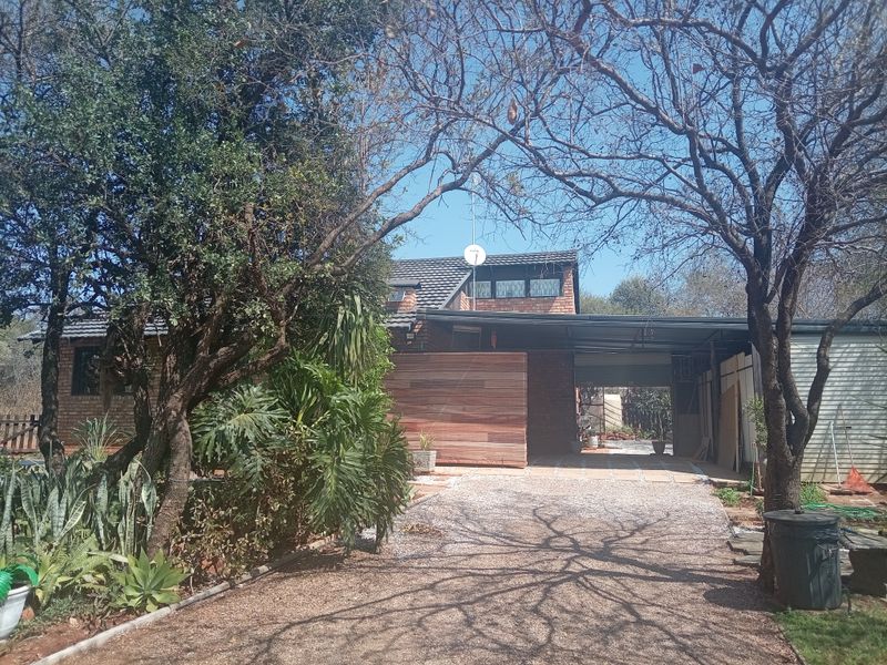 Front view of a brick house featuring a gravel driveway, carport, and mature trees in a residential setting.