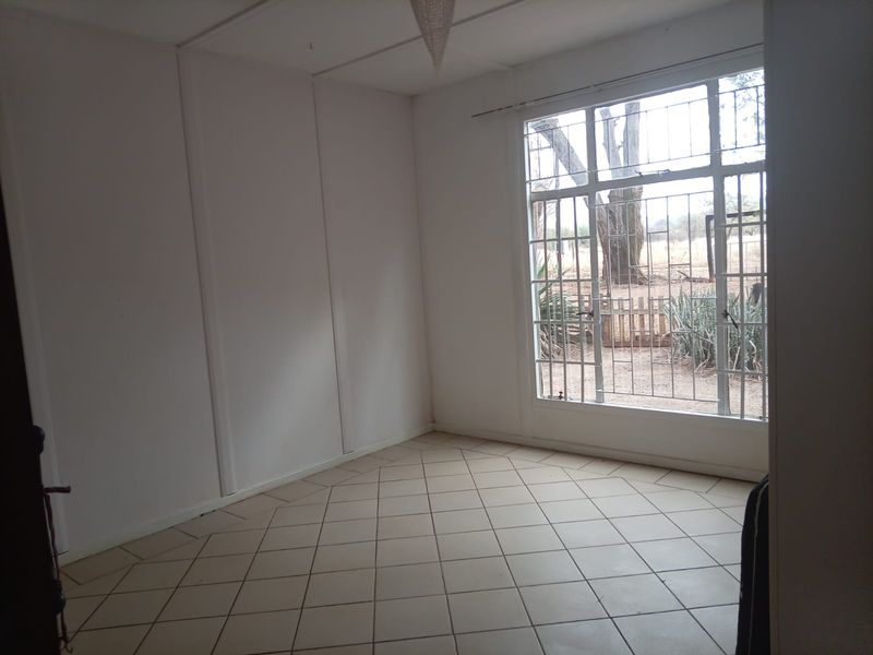 An empty living room featuring neutral tiled flooring and a large window with security bars, offering a bright and versatile space.