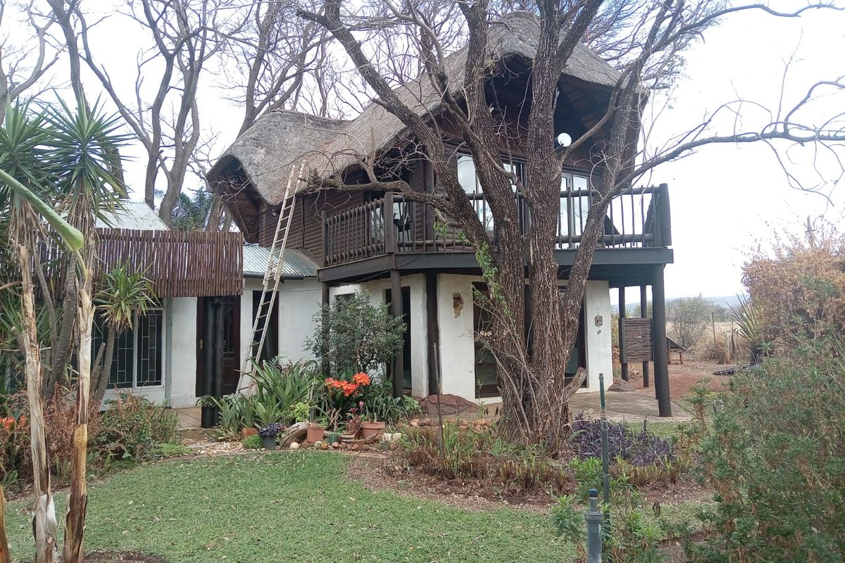 A two-story house featuring a traditional thatch roof, timber balcony, and sliding glass doors, set within a mature garden environment.