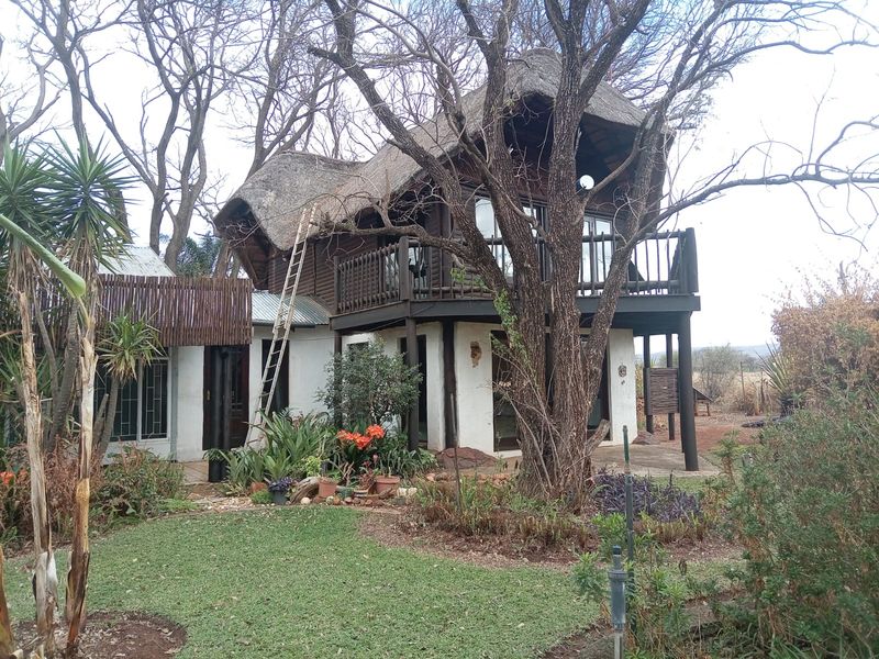 A two-story house featuring a traditional thatch roof, timber balcony, and sliding glass doors, set within a mature garden environment.