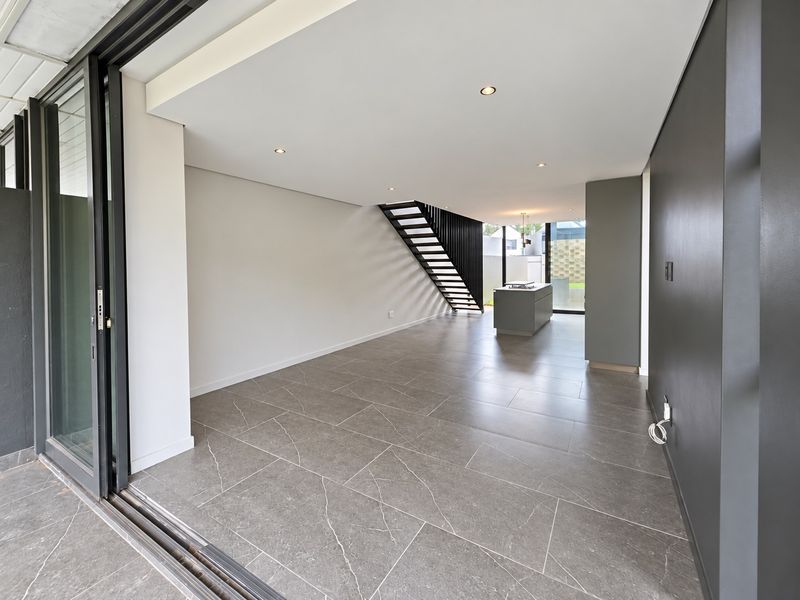 Modern open-plan living area featuring large format floor tiles, a sleek staircase, and a central kitchen island, seamlessly connecting to the outdoor patio.