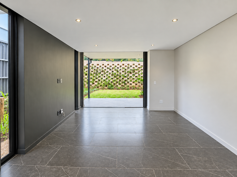 A bright, modern, and empty living area featuring large format floor tiles, recessed lighting, and seamless access to a private patio and garden.