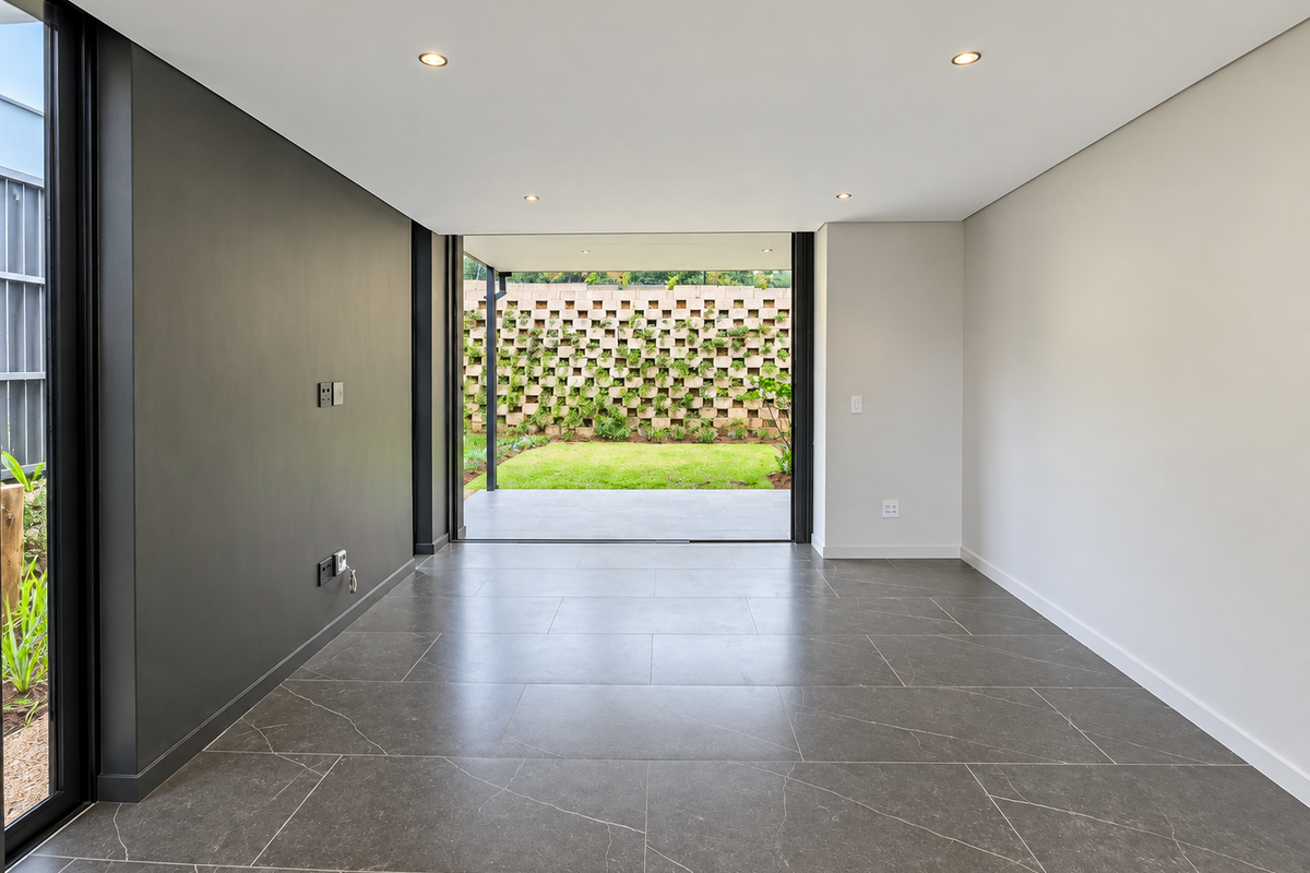 A bright, modern, and empty living area featuring large format floor tiles, recessed lighting, and seamless access to a private patio and garden.