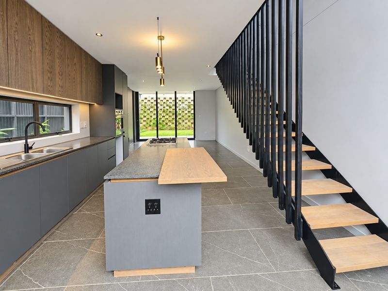 Modern open-plan kitchen featuring a central island, stone countertops, sleek cabinetry, and a striking staircase with timber treads and black metal balustrades.