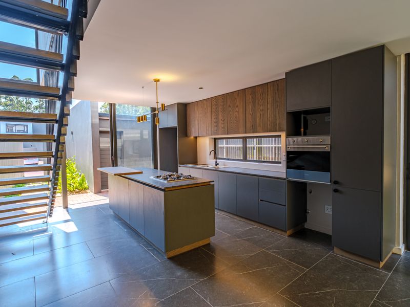 Modern kitchen featuring dark cabinetry, a central island with gas cooktop, built-in oven, and sleek stone countertops, adjacent to a striking floating staircase.