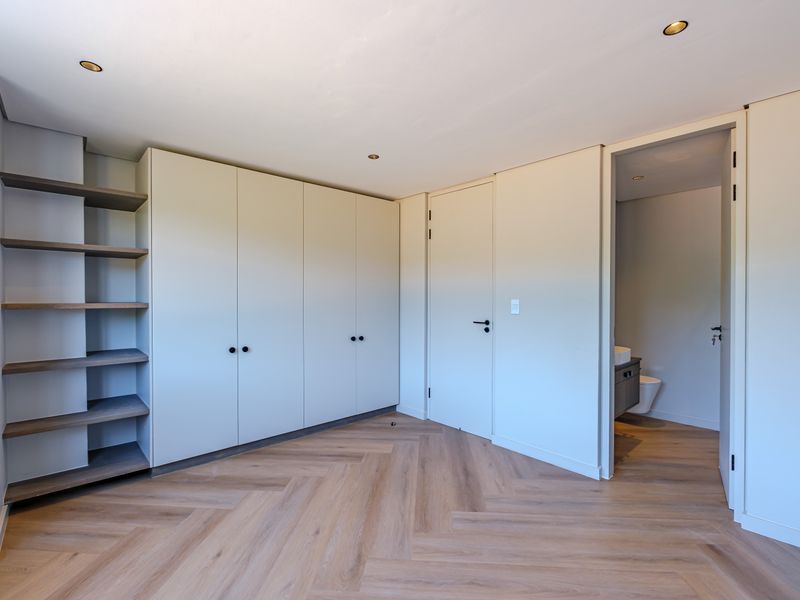 A modern, empty bedroom featuring light herringbone wood flooring, extensive built-in cabinetry with open shelving, and direct access to an en-suite bathroom.