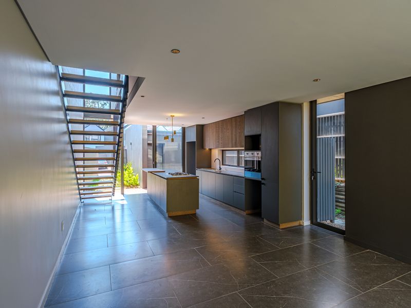 Modern open-plan kitchen featuring a central island, sleek cabinetry, built-in appliances, and a striking staircase, set against dark tiled flooring.