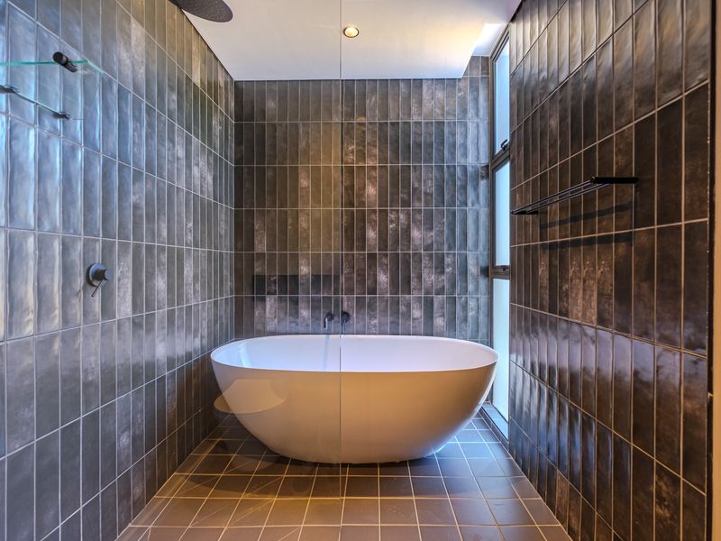 Modern bathroom featuring a sleek freestanding bathtub, floor-to-ceiling dark textured tiling, and a glass shower screen with contemporary fixtures.