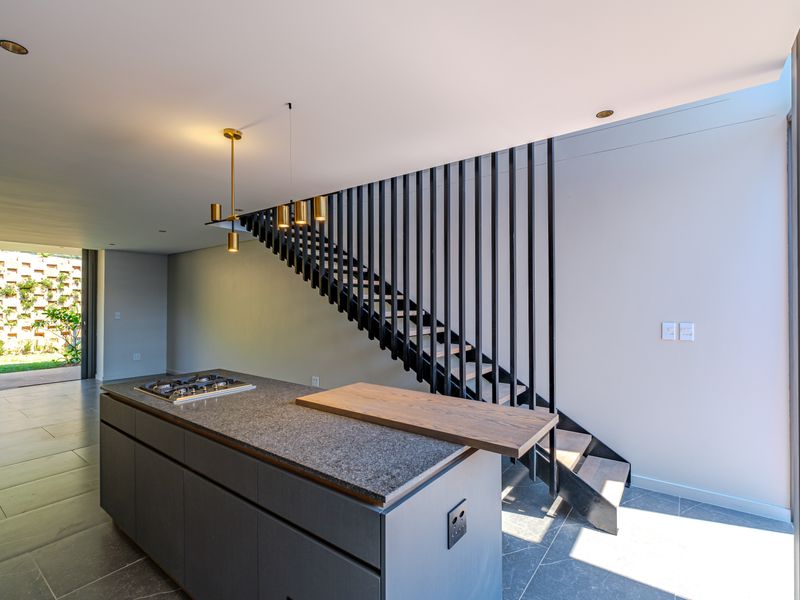Modern open-plan kitchen area featuring a central island with gas cooktop, stone countertop, and a striking architectural staircase with metal balustrade.