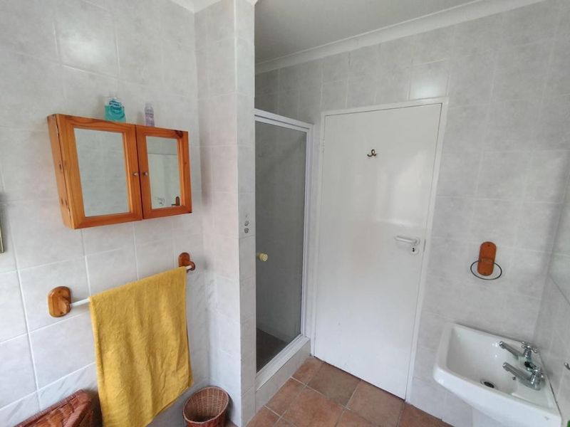 A functional bathroom featuring a shower enclosure, pedestal basin, and neutral wall tiling with terracotta-style floor tiles.