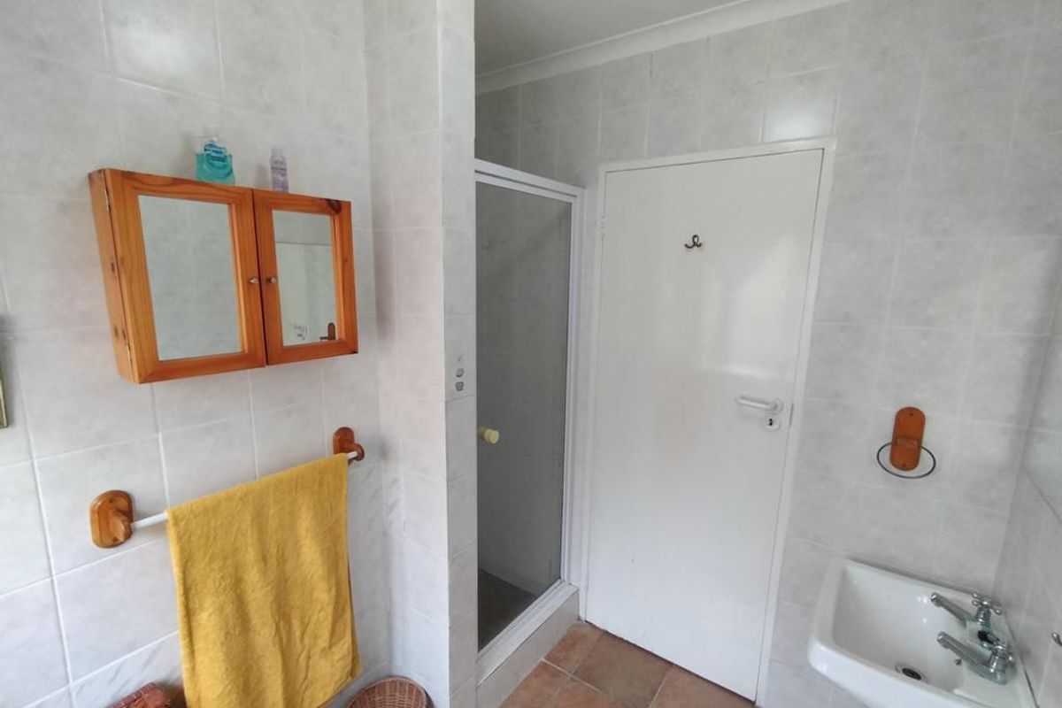 A functional bathroom featuring a shower enclosure, pedestal basin, and neutral wall tiling with terracotta-style floor tiles.