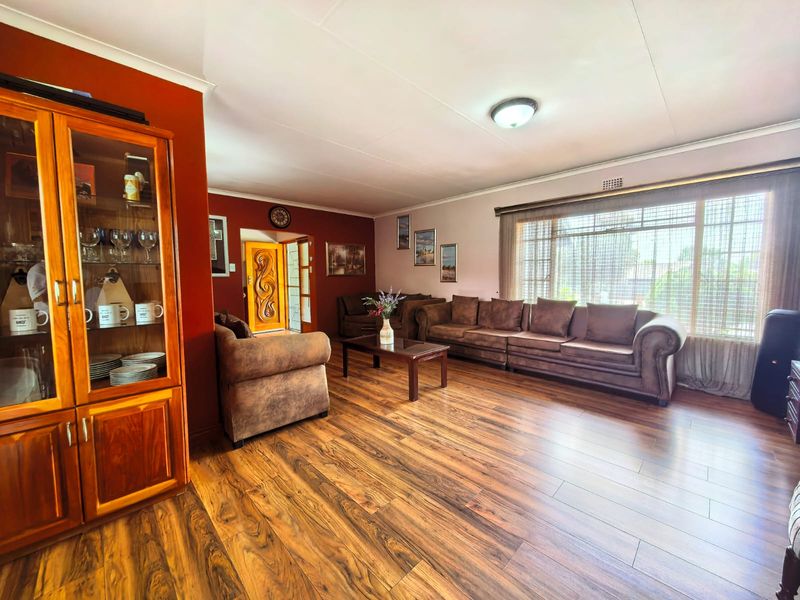 Spacious living room featuring warm-toned laminate flooring, large windows for natural light, and a decorative wooden entrance door.