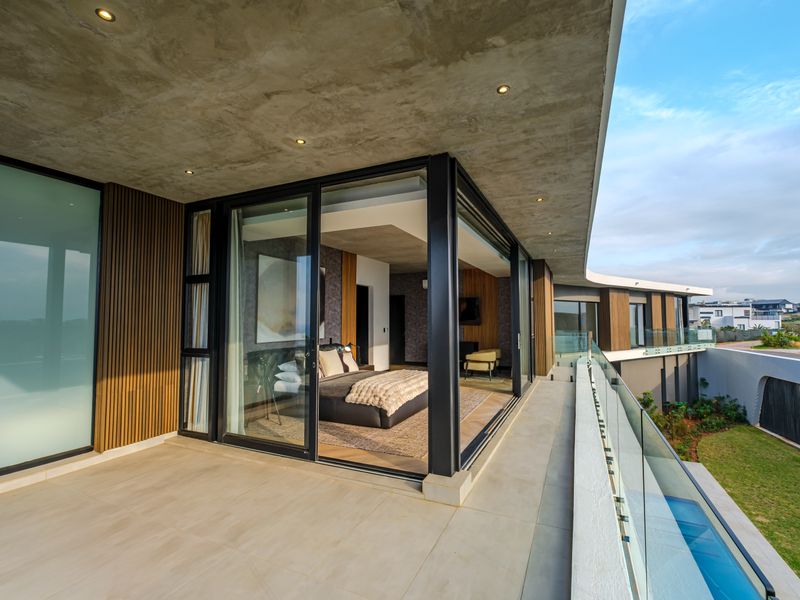 A modern covered balcony featuring a concrete ceiling, glass balustrade, and timber slat accents, providing seamless indoor-outdoor flow to the bedroom.