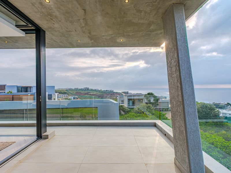 A modern covered balcony featuring tiled flooring, a glass balustrade, and expansive coastal views, seamlessly connected to the interior via sliding glass doors.