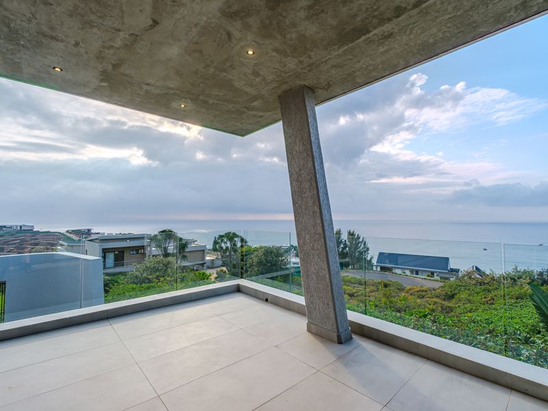 A modern, covered balcony featuring a glass balustrade, tiled flooring, and a concrete ceiling, offering expansive coastal and neighbourhood views.