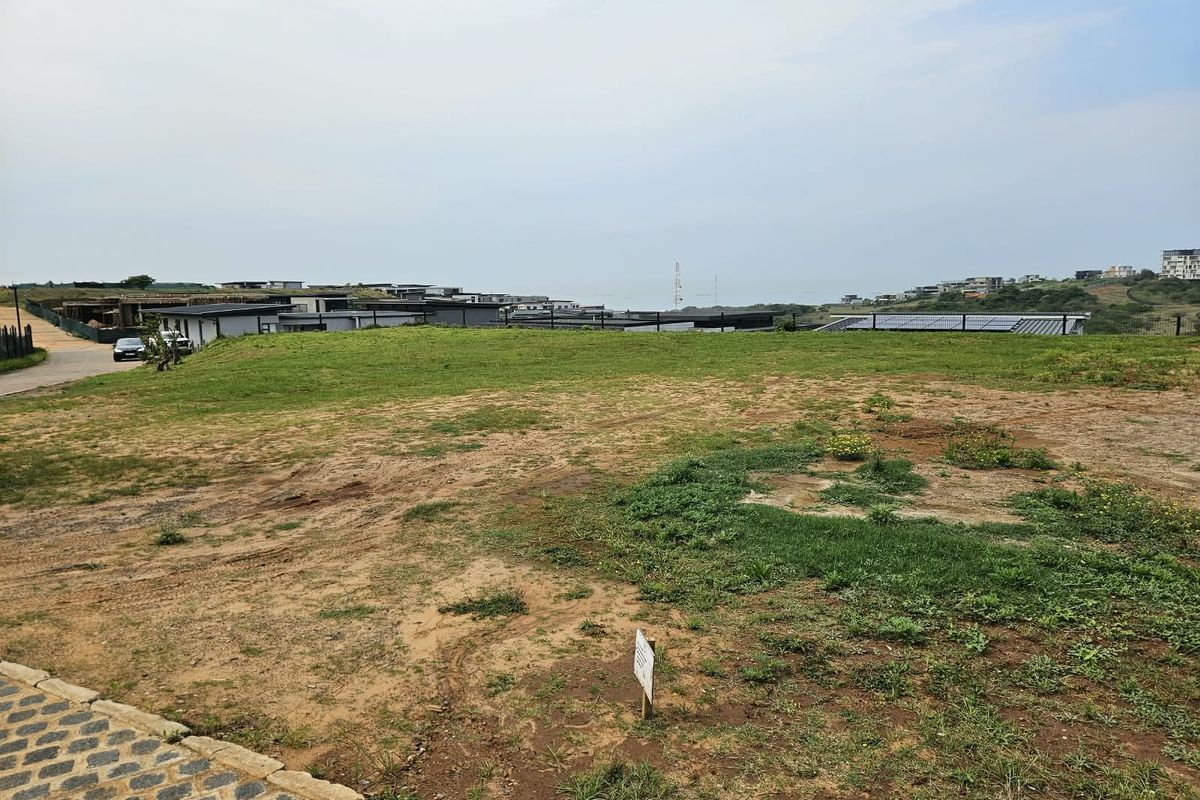 A vacant residential plot featuring a paved driveway, grassed terrain, and perimeter fencing, situated within a developed estate with distant coastal views.