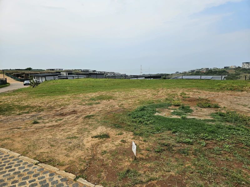 A vacant residential plot featuring a paved driveway, grassed terrain, and perimeter fencing, situated within a developed estate with distant coastal views.