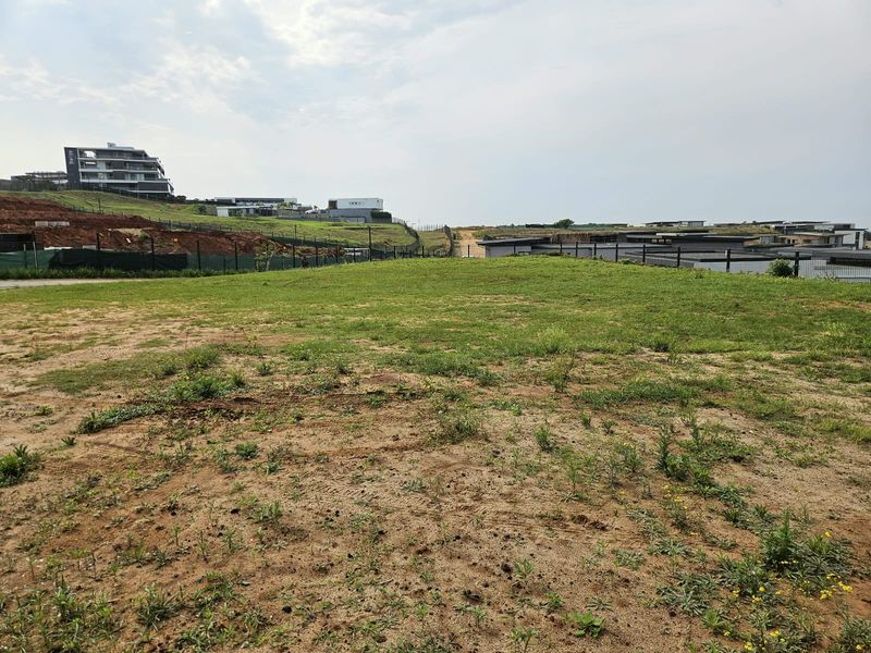 A vacant residential plot featuring grassed terrain and perimeter fencing, situated within a developed estate with surrounding modern residential properties.