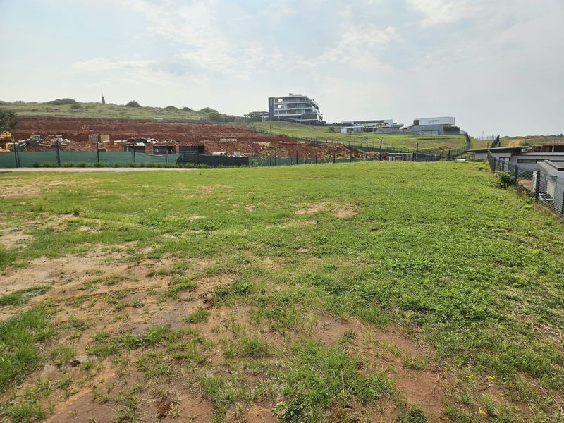 A view of a vacant residential plot featuring a grass lawn and perimeter fencing, situated within a developing estate.
