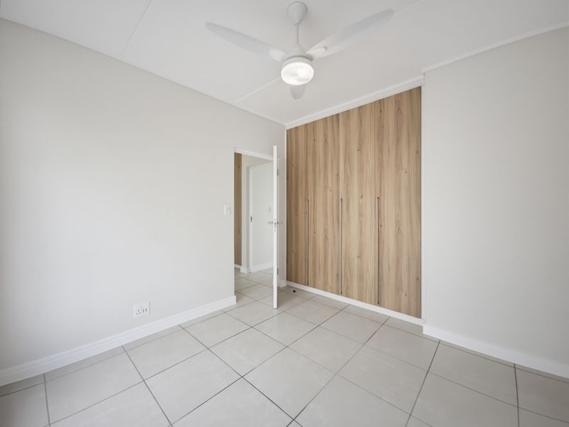 A bright, modern bedroom featuring neutral tiled flooring, a ceiling fan, and sleek wood-finish built-in cupboards.