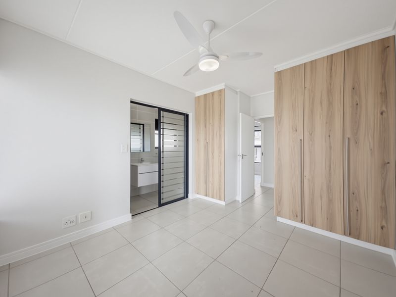 A bright, modern bedroom featuring neutral tiled flooring, built-in wood-finish cupboards, a ceiling fan, and direct access to an en-suite bathroom.