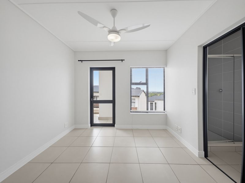 A bright, empty bedroom featuring neutral tiled flooring, a modern ceiling fan, and direct access to an outdoor balcony through a glass-panelled door.