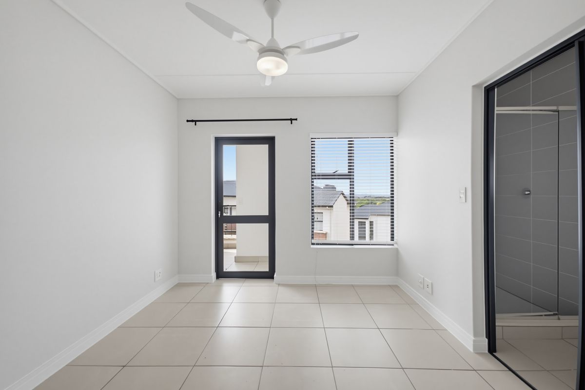 A bright, empty bedroom featuring neutral tiled flooring, a modern ceiling fan, and direct access to an outdoor balcony through a glass-panelled door.