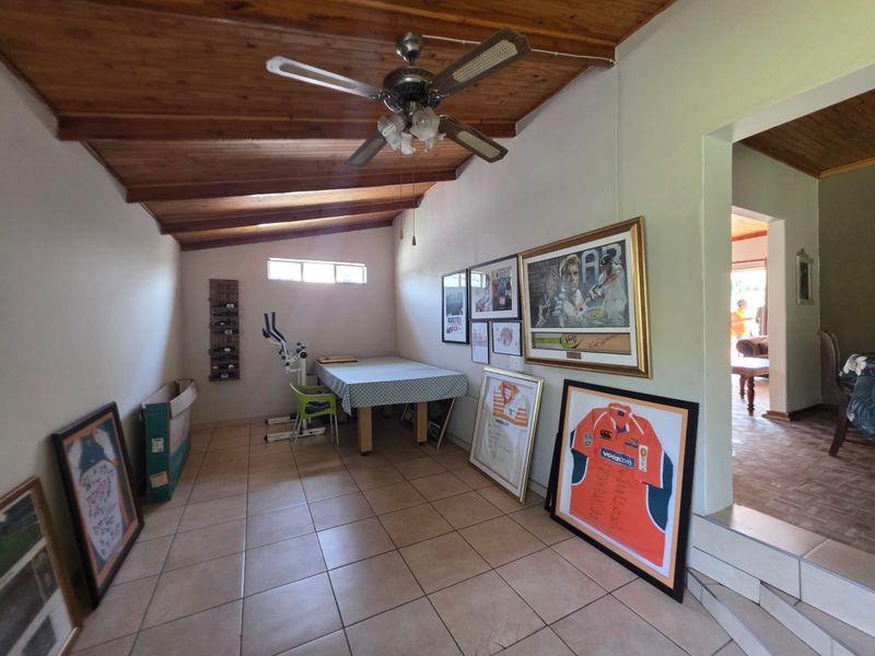 A living area featuring an exposed timber raked ceiling, ceiling fan, and tiled flooring, with a view into an adjacent room.