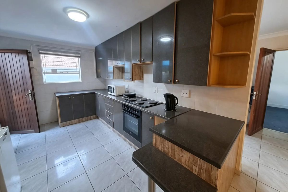 A functional kitchen featuring dark cabinetry, stone countertops, a built-in oven with electric hob, and a breakfast bar, finished with neutral floor tiles.