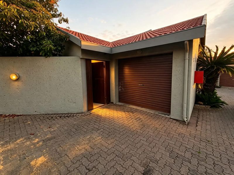 A well-maintained townhouse garage featuring a roller door, tiled roof, and paved driveway, illuminated by exterior wall lighting.