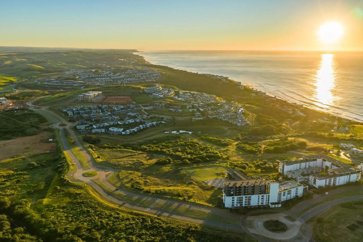 An expansive aerial view of a coastal residential estate featuring modern multi-story buildings, established road infrastructure, and scenic ocean views at sunset.