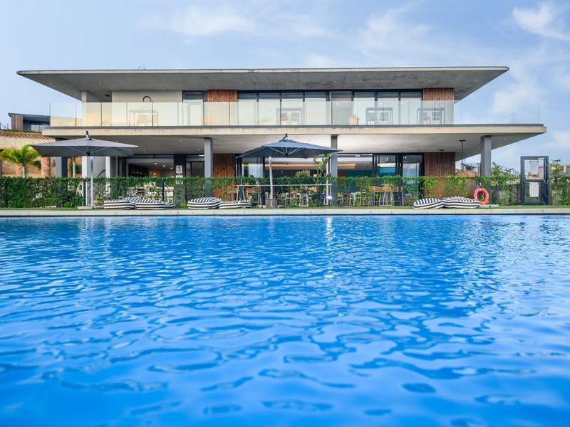 A modern two-story building featuring a large swimming pool in the foreground, glass balustrades, and a spacious covered patio area.