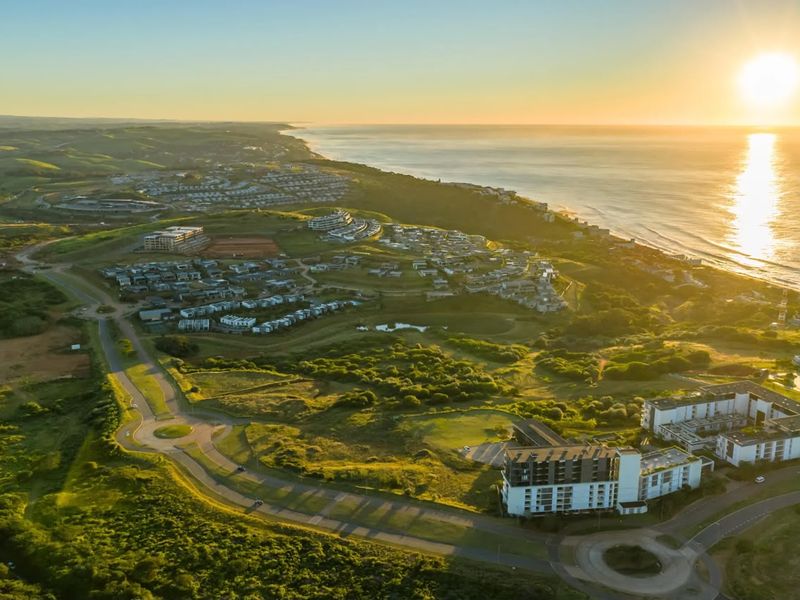 An expansive aerial view of a coastal residential estate featuring modern multi-story buildings, established road infrastructure, and scenic ocean views at sunset.