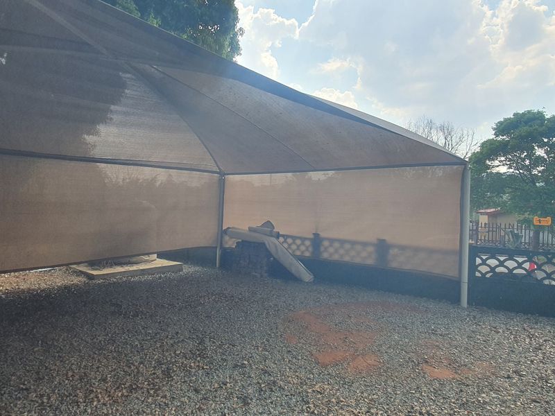 An outdoor carport area featuring a shade cloth structure and gravel driveway, enclosed by a perimeter boundary wall.