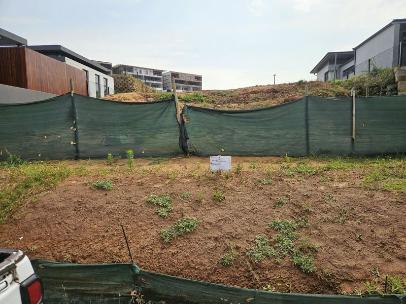 A vacant residential plot featuring sloped terrain, enclosed by temporary green shade cloth fencing, situated within a developed neighborhood.