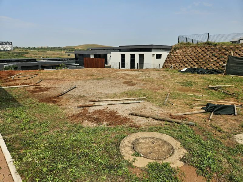A vacant residential plot featuring a retaining wall, manhole cover, and perimeter fencing, offering a clear view of the surrounding estate.