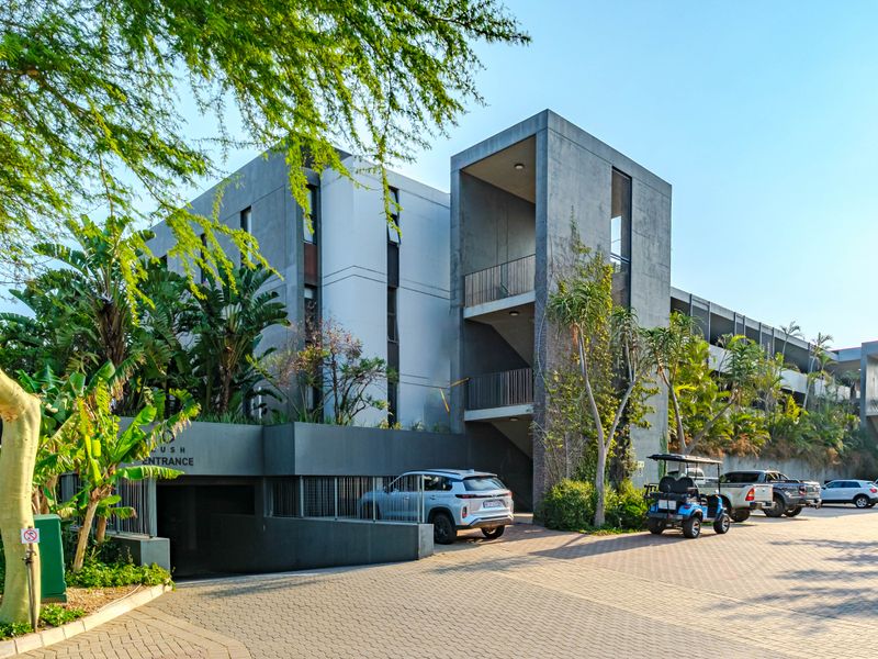 Modern apartment building exterior featuring a concrete facade, paved driveway, and secure underground parking entrance with established landscaping.
