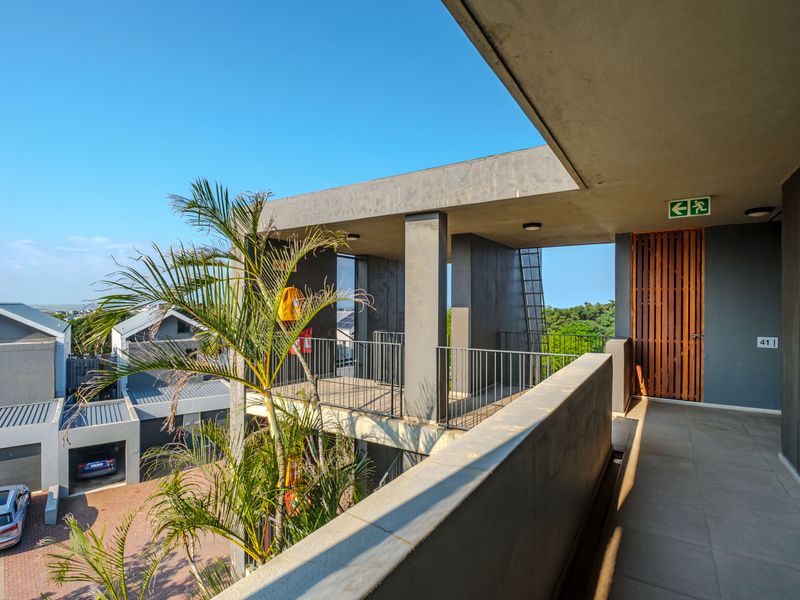 A modern, open-air apartment balcony featuring concrete construction, metal safety railings, and a timber-slat entrance door with recessed lighting.
