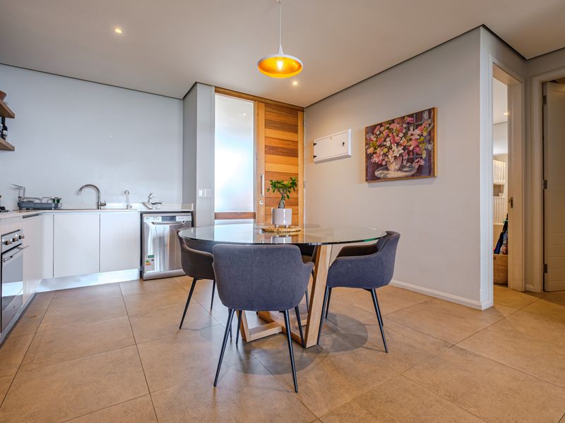 Modern open-plan kitchen and dining area featuring sleek white cabinetry, integrated appliances, tiled flooring, and a warm timber door entrance.